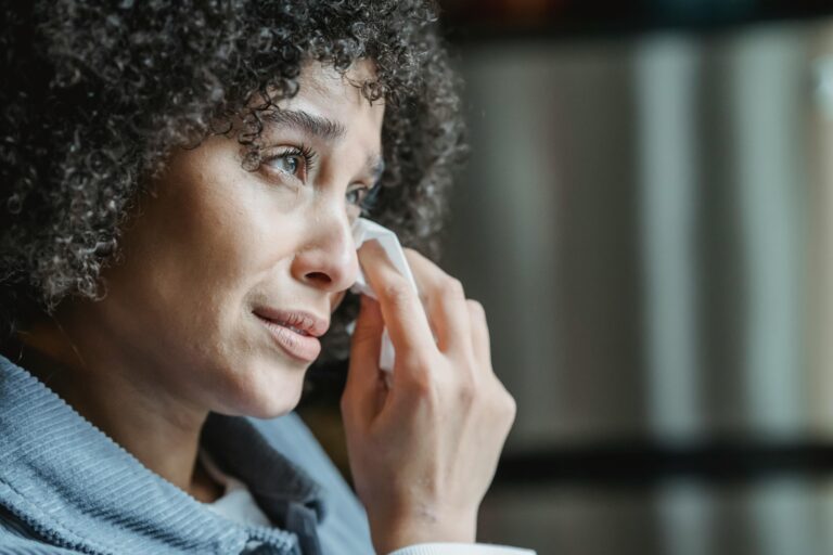 Side view of crop African American sorrowful lady with curly hair wiping tears with napkin while crying near window at home
